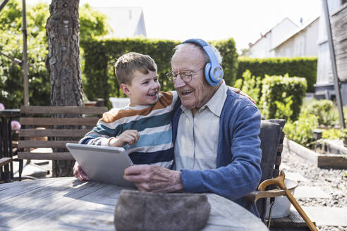 Glücklicher Junge mit Großvater, der drahtlose Kopfhörer trägt und einen Tablet-PC benutzt - UUF29021
