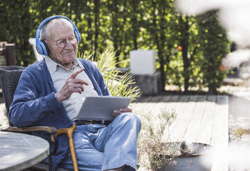 Nachdenklicher älterer Mann mit drahtlosen Kopfhörern, der mit einem Tablet-PC sitzt - UUF29033