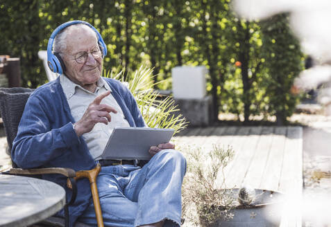 Nachdenklicher älterer Mann mit drahtlosen Kopfhörern, der mit einem Tablet-PC sitzt - UUF29033