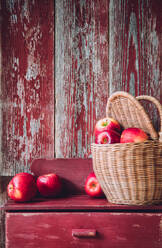 Wicker basket filled with fresh ripe red apples placed on shabby timber cupboard against wooden wall in countryside house during harvest season - ADSF44355