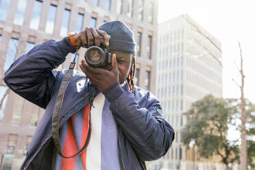Black male photographer with professional photo camera standing against building and taking picture in city street - ADSF44361