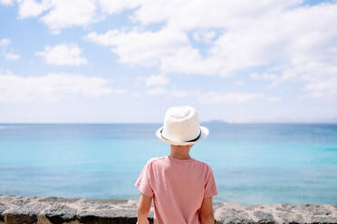 Back view boy in hat standing in daylight while looking away against blurred sea and blue sky with clouds - ADSF44375