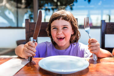 Cute hungry preschool girl in purple t shirt sitting at wooden table with opened mouth and silverware in hands while waiting for order in cafe - ADSF44379