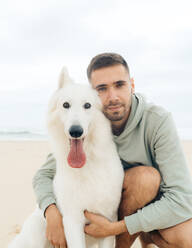 Content young male owner in hoodie hugging fluffy white Shepherd dog and looking at camera on sandy beach in Vieux Boucau les Bains - ADSF44382