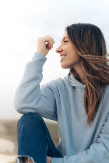 Positive young female in blue hoodie and jeans with long brown hair looking away and smiling while sitting on sandy beach in Vieux Boucau les Bains - ADSF44383