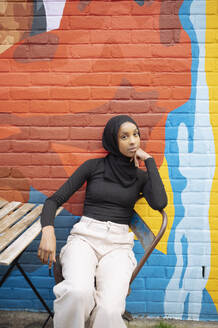 Young woman wearing hijab sitting against mural - ISF26147