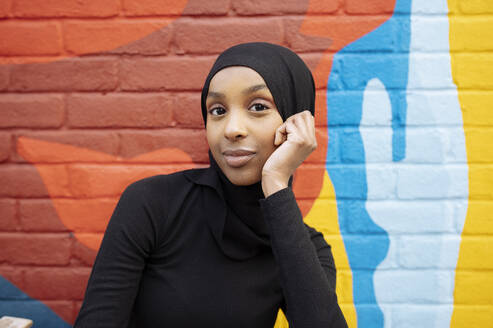 Portrait of young woman wearing hijab sitting against mural - ISF26148