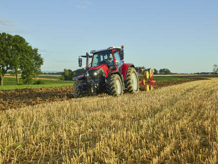 Landwirt mit Traktor und Pflügen Land in der Nähe von trockenem Gras bei Sonnenaufgang - CVF02447