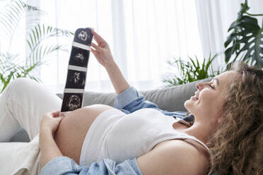 Pregnant woman looking at ultrasound scans lying on couch at home - AAZF00726