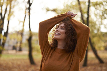 Young redhead woman with eyes closed and arms raised at autumn park - ABIF02041