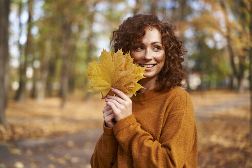 Lächelnde rothaarige Frau mit Ahornblättern im Herbstpark - ABIF02043