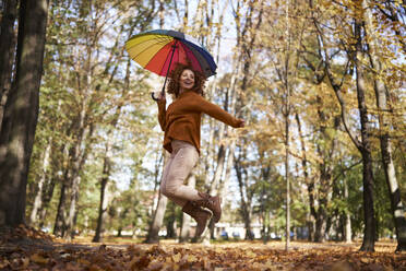 Smiling redhead woman jumping with colorful umbrella at autumn park - ABIF02052