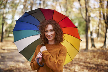 Lächelnde rothaarige Frau hält bunten Regenschirm im Herbst Park - ABIF02053