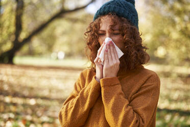 Redhead woman blowing nose with facial tissue at autumn park - ABIF02063