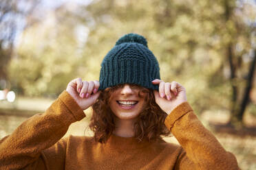 Cheerful redhead woman covering eyes with knit hat at autumn park - ABIF02064