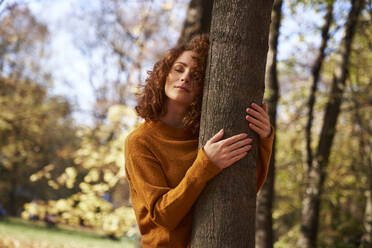 Redhead woman with eyes closed embracing tree at autumn park - ABIF02070