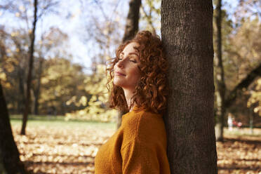 Rothaarige Frau mit geschlossenen Augen lehnt an einem Baum im Herbstpark - ABIF02072