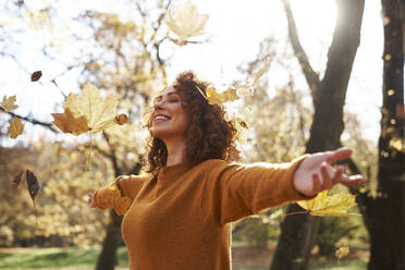 Autumn leaves falling on redhead woman with arms outstretched at park - ABIF02073