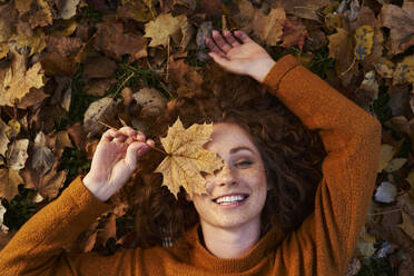Smiling redhead woman holding maple leaf and lying on ground at park - ABIF02075