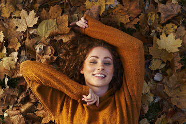 Smiling redhead woman lying on autumn leaves at park - ABIF02076
