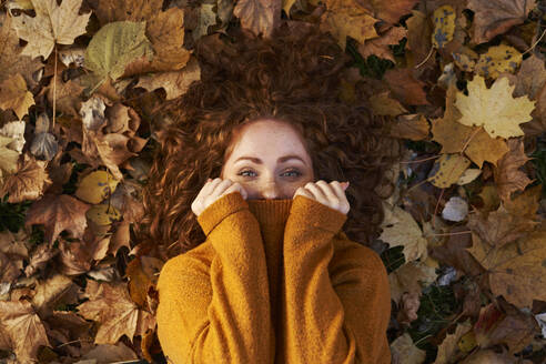 Redhead woman covering face with sweater lying on maple leaves at park - ABIF02077