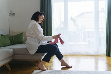 Smiling pregnant woman with baby clothes sitting on sofa in living room at home - NJAF00401