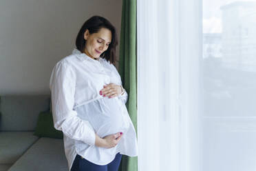 Lächelnde schwangere Frau, die zu Hause am Fenster steht - NJAF00407