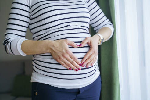 Schwangere Frau macht Herzform auf dem Bauch und steht am Fenster zu Hause - NJAF00408