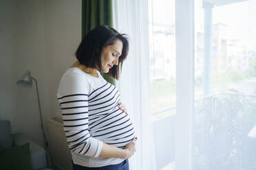 Lächelnde schwangere Frau, die ihren Bauch berührt und am Fenster steht - NJAF00409