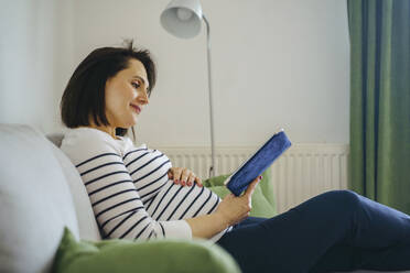 Smiling woman sitting on sofa and reading book at home - NJAF00410