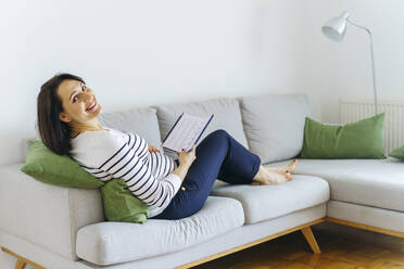 Happy pregnant woman sitting on sofa with book in living room at home - NJAF00413