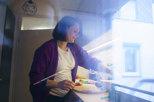 Lächelnde schwangere Frau, die in der Küche Essen zubereitet, gesehen durch Glas - NJAF00416