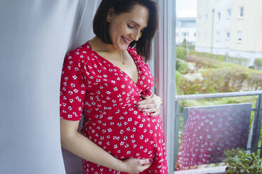 Glückliche schwangere Frau, die ihren Bauch berührt und zu Hause am Fenster steht - NJAF00423