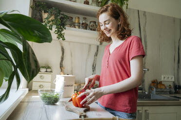 Happy young woman cutting red bell pepper in kitchen at home - OSF01755