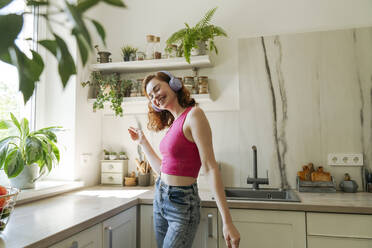 Glückliche Frau mit drahtlosen Kopfhörern tanzt in der Küche zu Hause - OSF01761