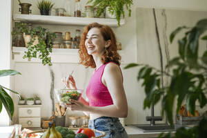 Happy young woman with bowl of salad standing in kitchen at home - OSF01771