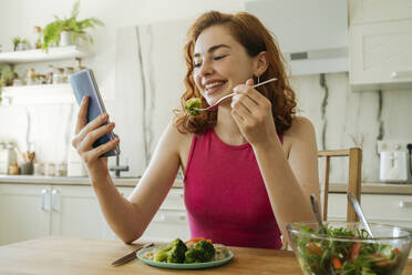 Glückliche junge Frau, die ein Smartphone benutzt und am Esstisch Salat isst - OSF01772