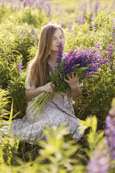 Woman smelling purple lupine flowers sitting in field - ONAF00559