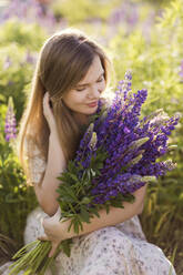 Smiling woman holding bunch of purple lupine flowers in field - ONAF00560