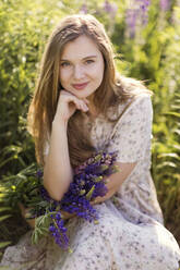 Smiling woman with hand on chin and purple lupine flowers sitting in field - ONAF00563