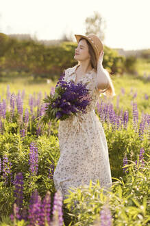 Lächelnde Frau mit Hut, stehend mit lila Lupinenblüten - ONAF00565