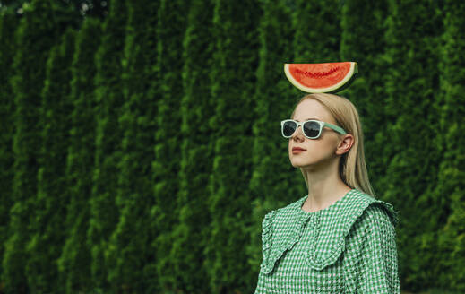 Woman wearing sunglasses standing with watermelon on head in garden - VSNF01092