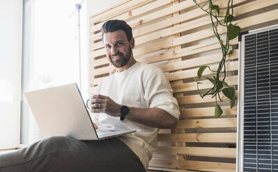 Glücklicher Geschäftsmann mit Laptop und Kaffeetasse zu Hause - UUF29040