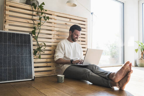 Glücklicher Geschäftsmann, der an einem Sonnenkollektor sitzt und einen Laptop zu Hause benutzt - UUF29043