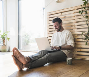 Happy businessman using laptop on floor at home - UUF29045