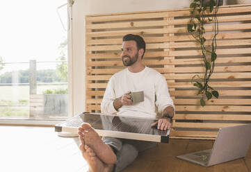 Glücklicher Geschäftsmann mit Kaffeetasse und Solarpanel zu Hause sitzend - UUF29050