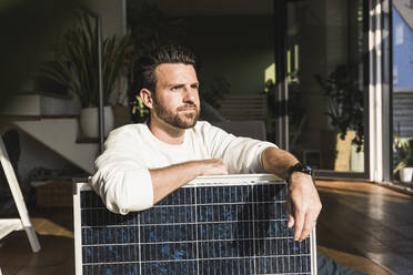 Nachdenklicher Geschäftsmann sitzt mit Solarpanel zu Hause - UUF29056