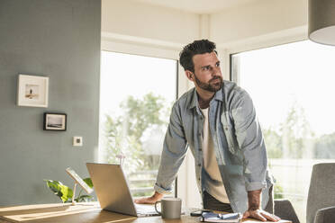 Thoughtful businessman leaning on desk in home office - UUF29073