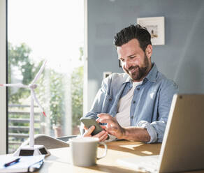 Happy businessman using smart phone at desk - UUF29077