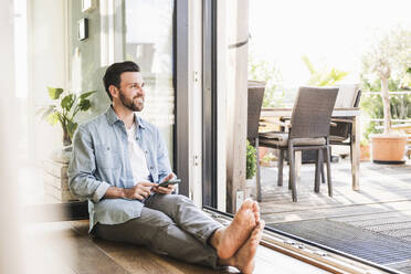 Thoughtful businessman sitting with smart phone on floor - UUF29097